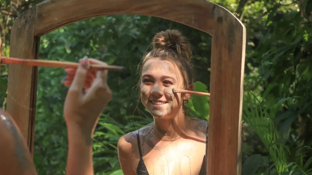 Woman applying makeup in mirror outdoors with greenery around.