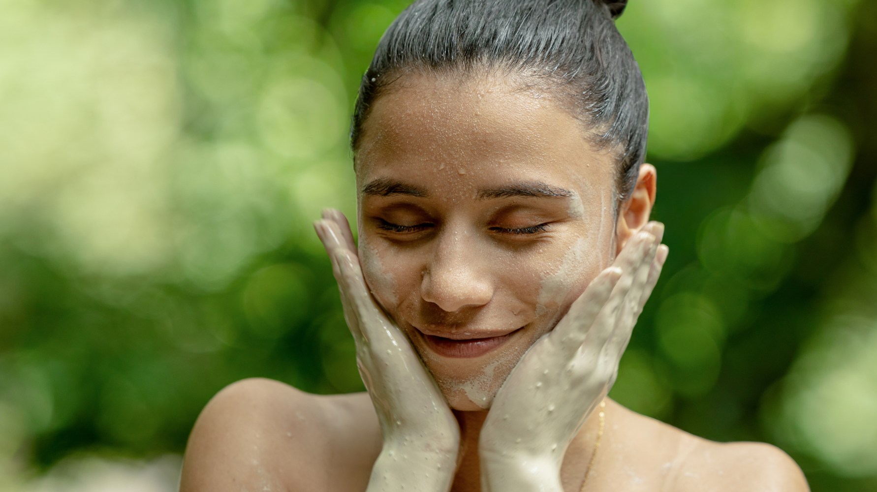 Mujer con mascarilla de barro en la cara sonriendo y tocándose las mejillas al aire libre.