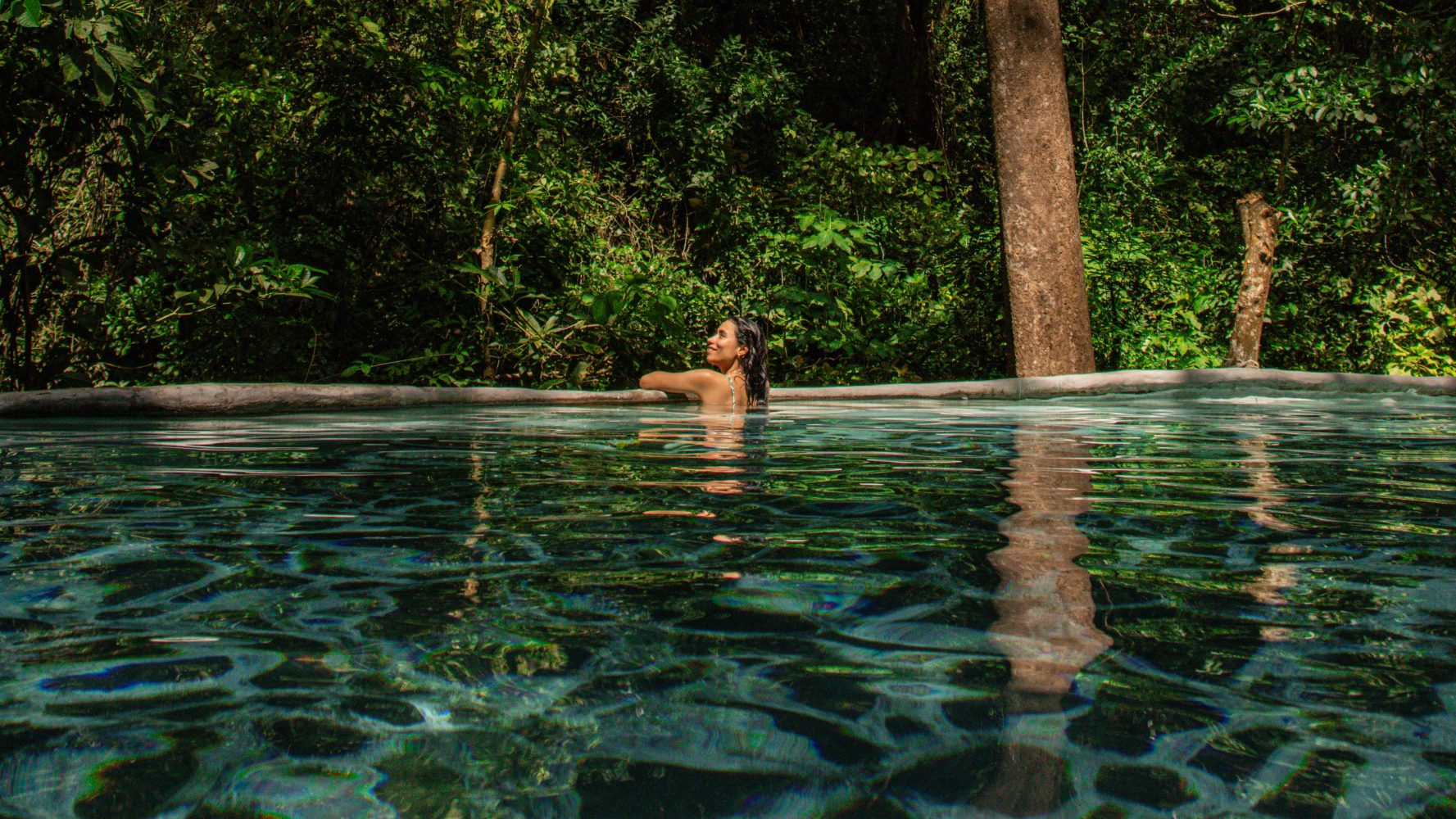 Persona nadando en una piscina clara rodeada de exuberantes árboles verdes.