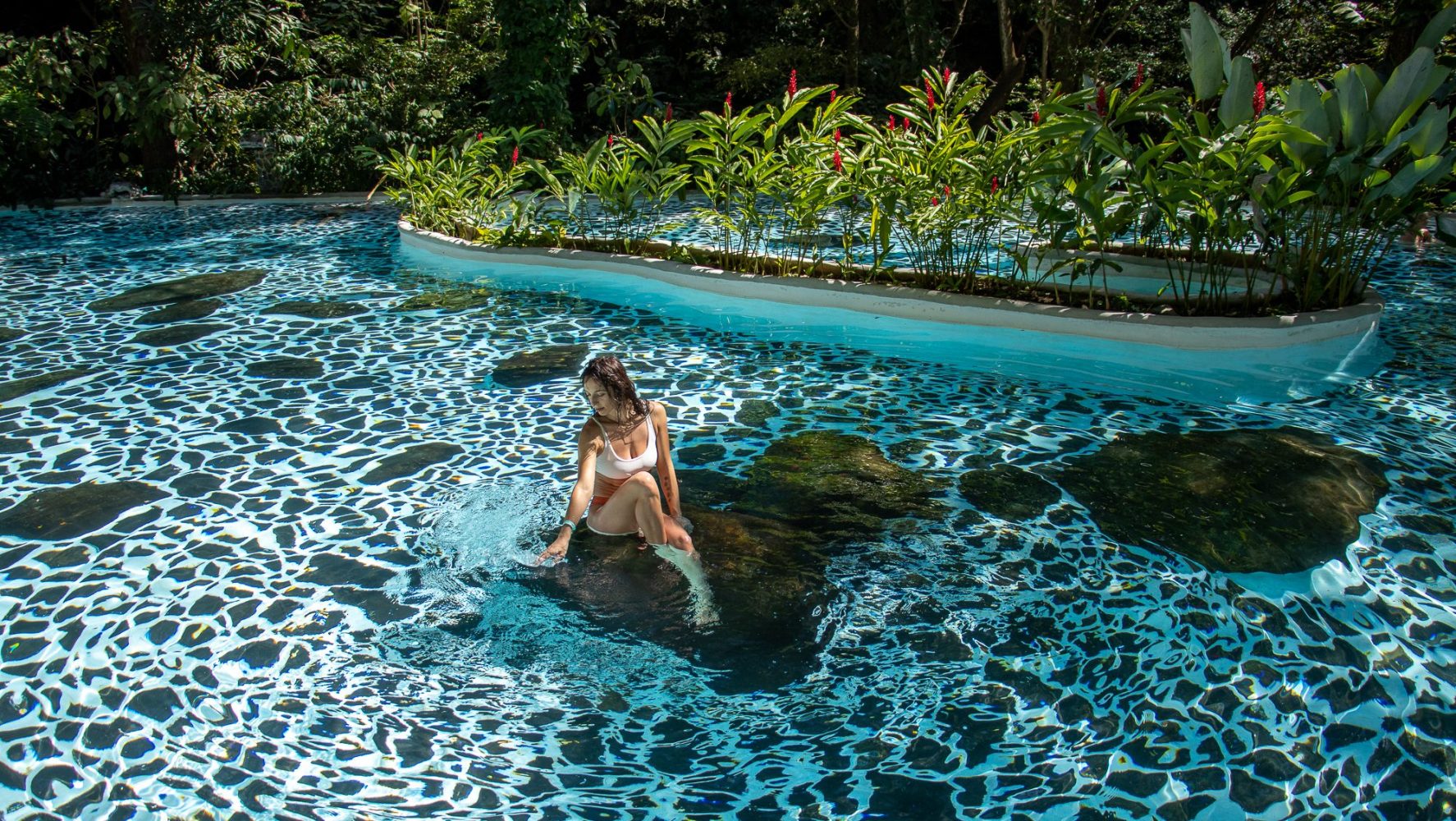 Mujer en bikini blanco sentada en una roca en una piscina estampada con plantas exuberantes alrededor.