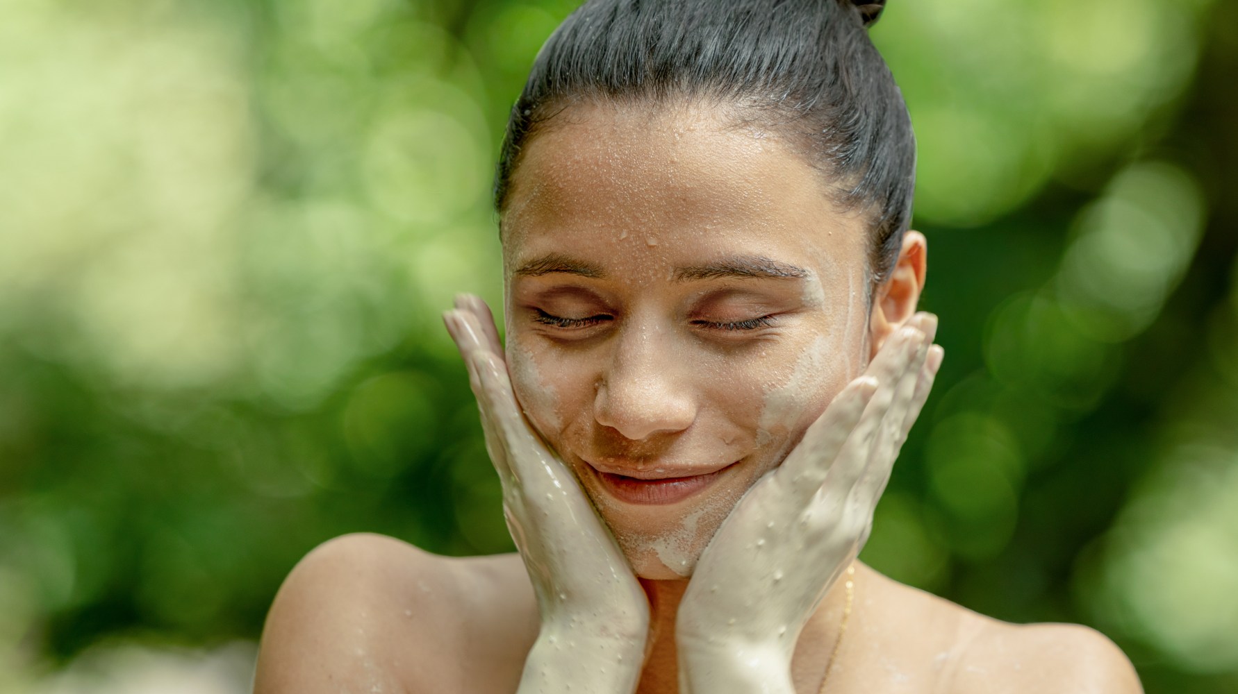 Persona con mascarilla de arcilla en la cara, sonriendo con los ojos cerrados, manos en las mejillas, fondo verde.