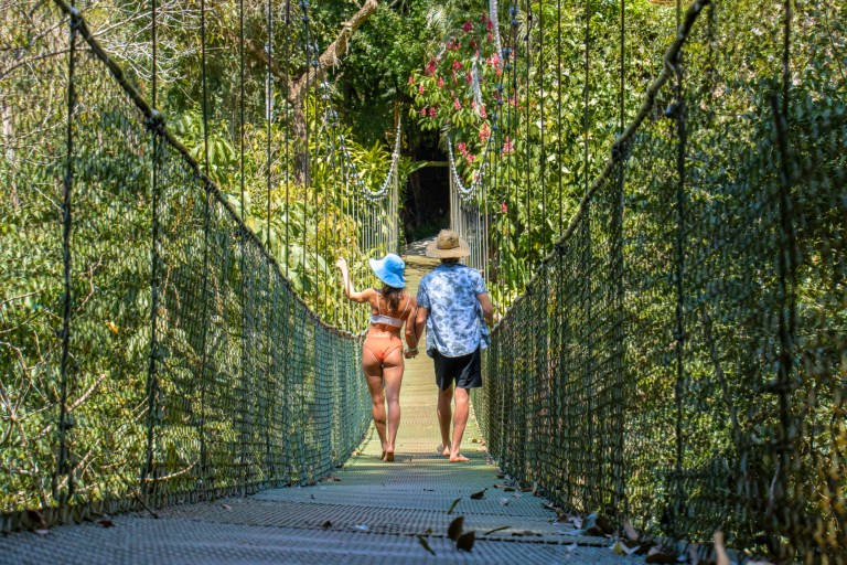 Two people in hats walk on a suspension bridge surrounded by lush greenery.