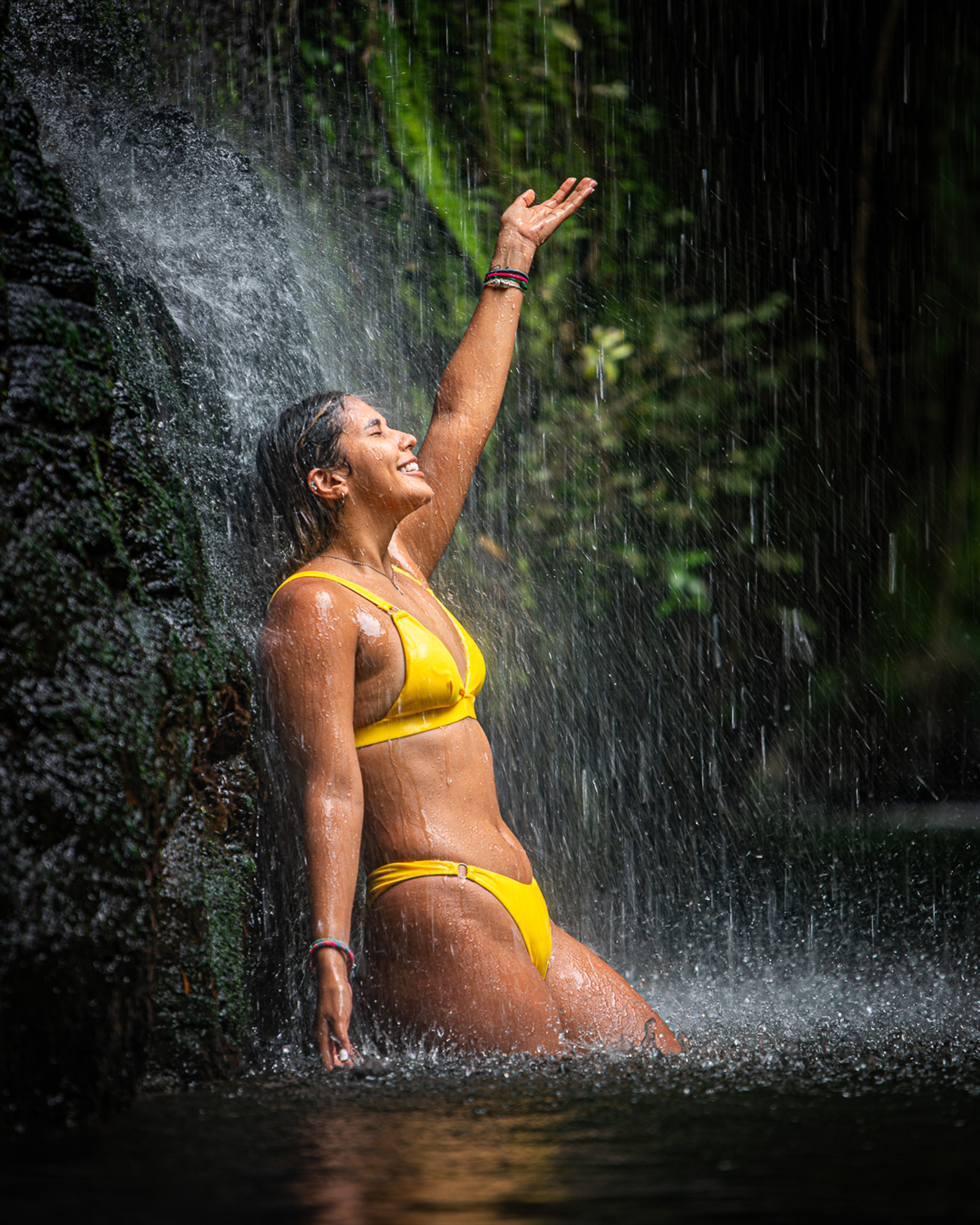 Woman in yellow bikini enjoying waterfall, reaching up with one arm, surrounded by greenery.