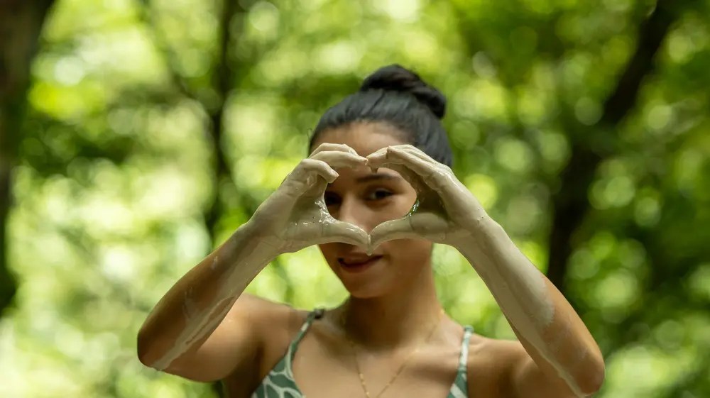 Person forms heart shape with hands, green background.