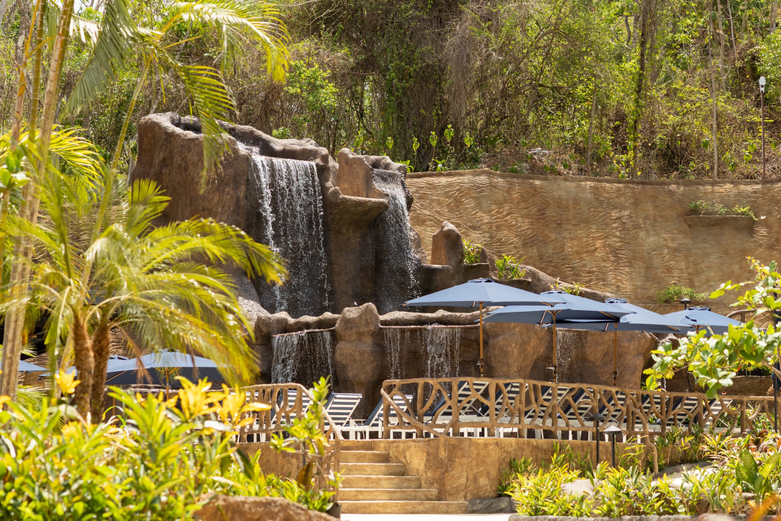 Waterfall with rocks, palm trees, and lounge chairs under umbrellas.