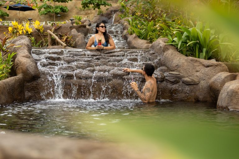 Two people relax in a stone-lined waterfall pool surrounded by lush greenery.