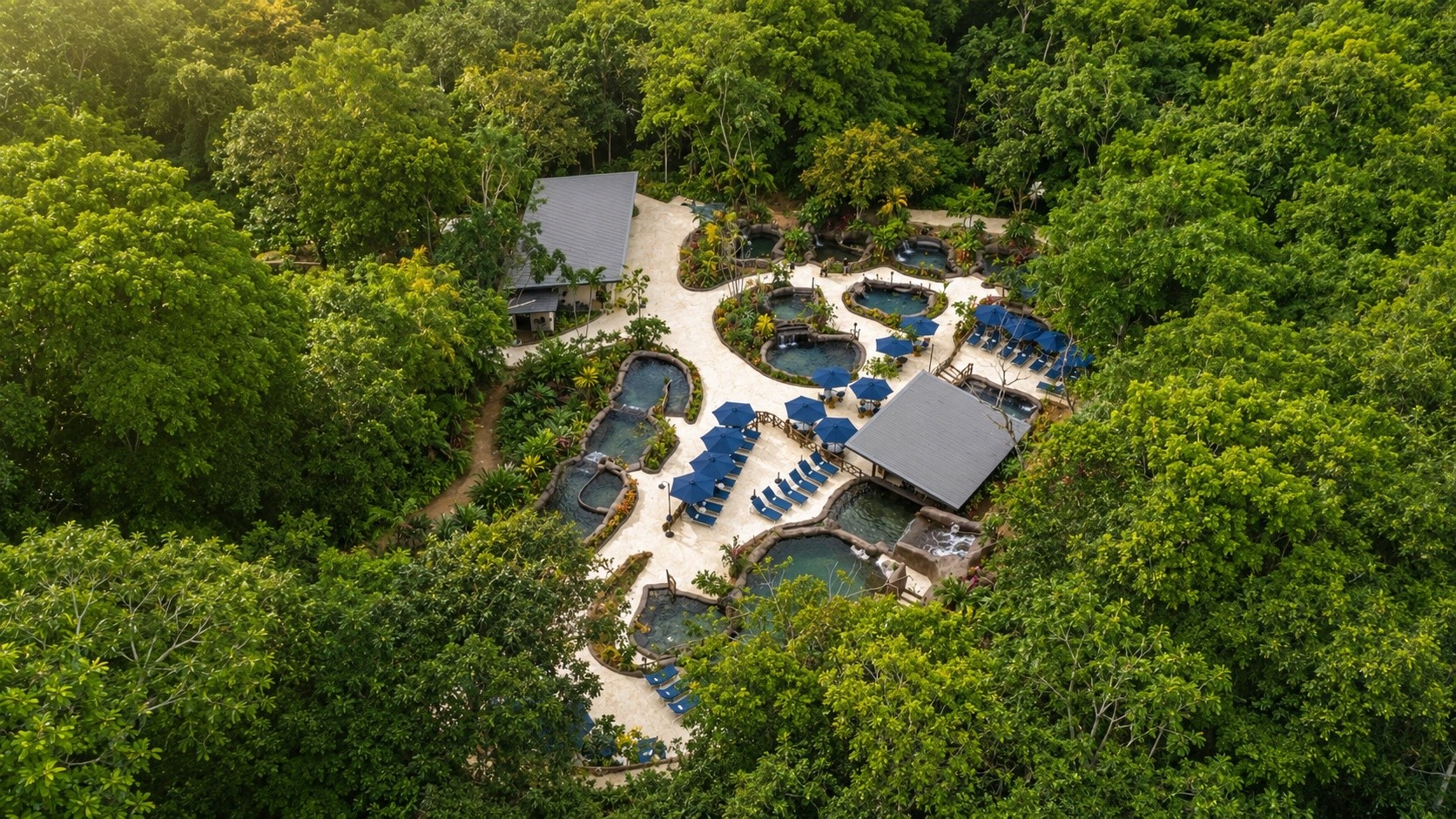 Aerial view of a resort surrounded by dense green forest with pools and blue umbrellas.